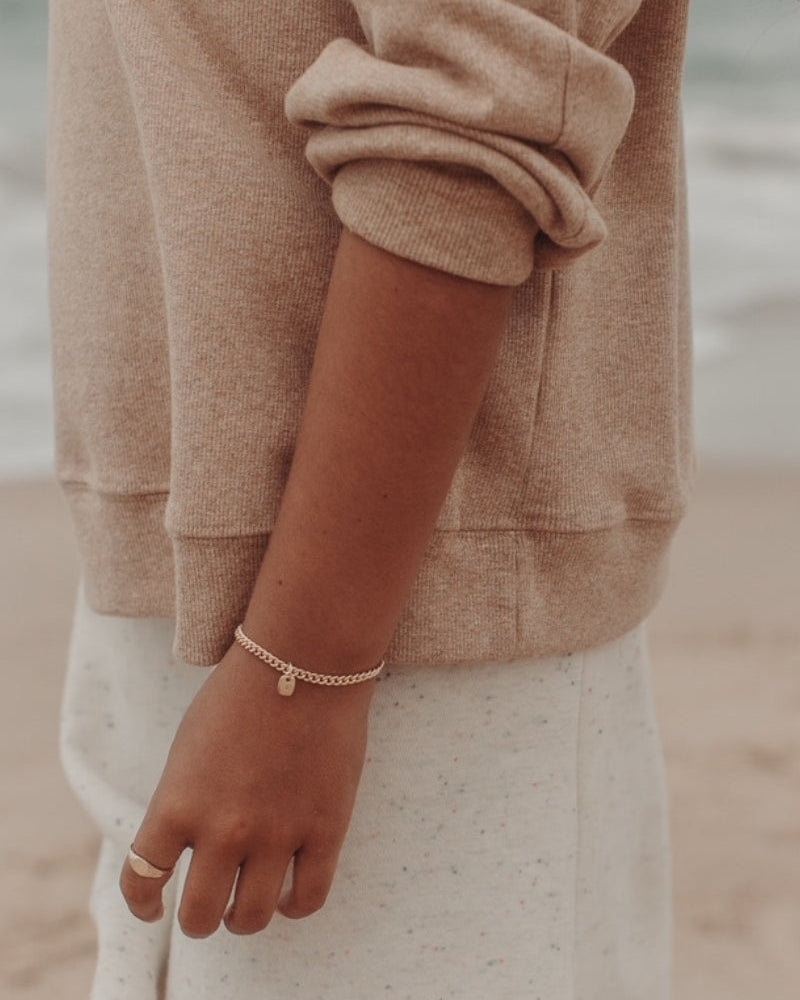 A close-up photo of a woman’s wrist at the beach featuring a gold Cuban Link Chain with a minimalist solid gold rectangular pendant and a matching signet ring, styled in fine jewellery fashion by Bluebird Co