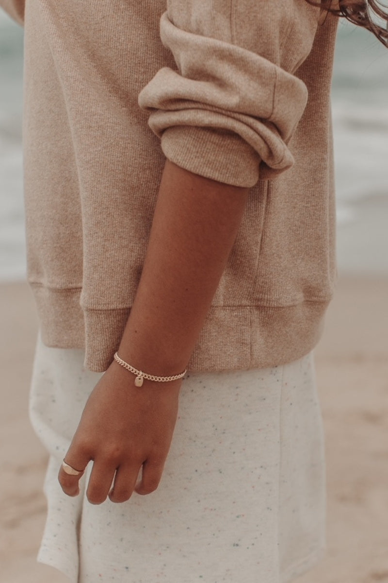 A close-up photo of a woman’s wrist at the beach featuring a gold Cuban Link Chain with a minimalist solid gold rectangular pendant and a matching signet ring, styled in fine jewellery fashion by Bluebird Co