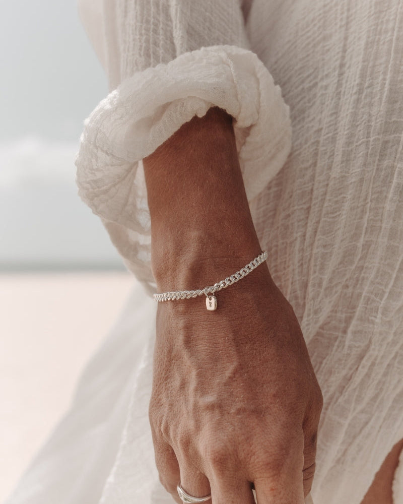 A close-up photo of a woman’s wrist at the beach featuring a Cuban Link Chain with a minimalist rectangular pendant and a matching signet ring, styled in fine jewellery fashion by Bluebird Co