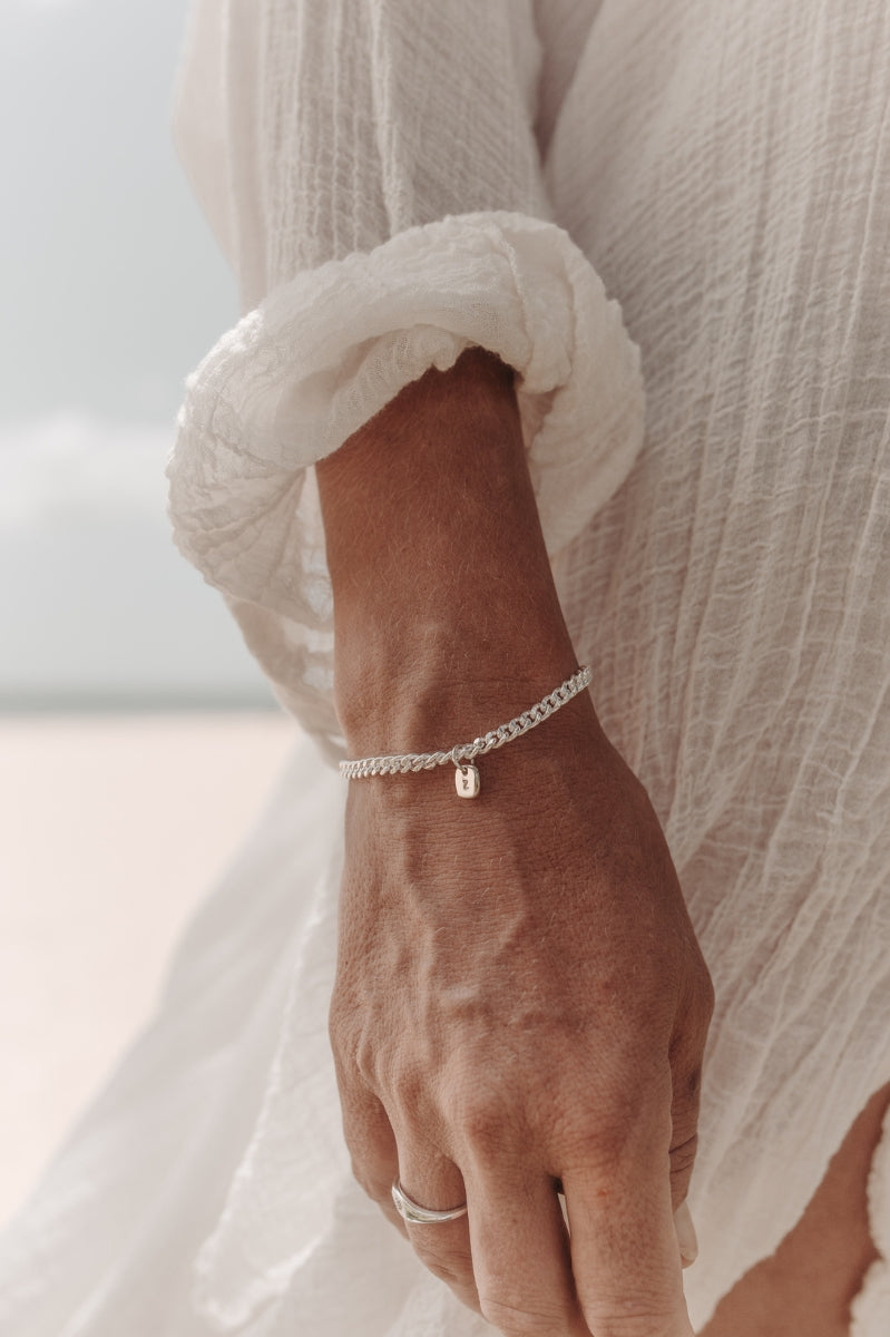 A close-up photo of a woman’s wrist at the beach featuring a Cuban Link Chain with a minimalist rectangular pendant and a matching signet ring, styled in fine jewellery fashion by Bluebird Co