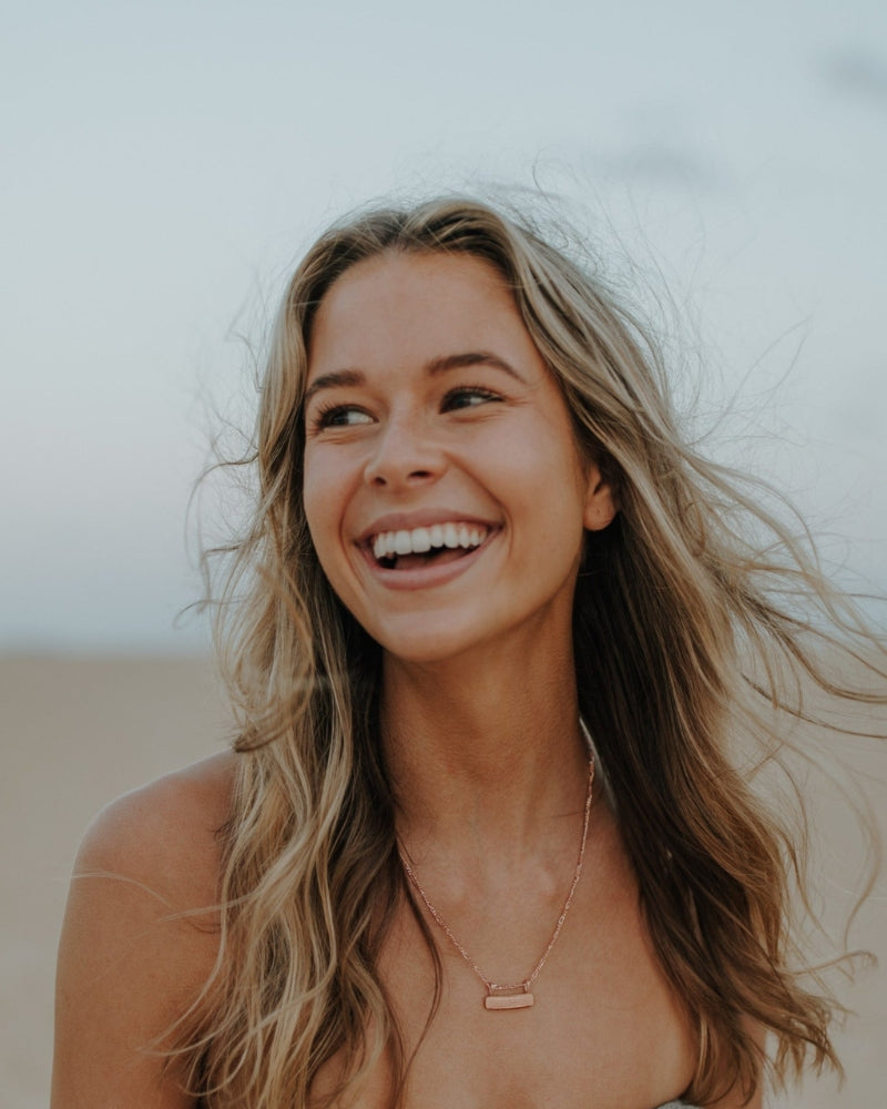 Rose gold Signature Bar Necklace shown on model laughing at the beach. Personal and stylish jewellery for everyday wear.