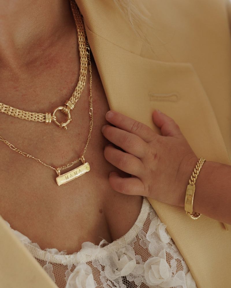 A mother is wearing the Signature Bar necklace in gold vermeil with the word 'MAMA' engraved on the front of it. It is layered with the Signature Statement Necklace and matches her daughter's Signature Bracelet.