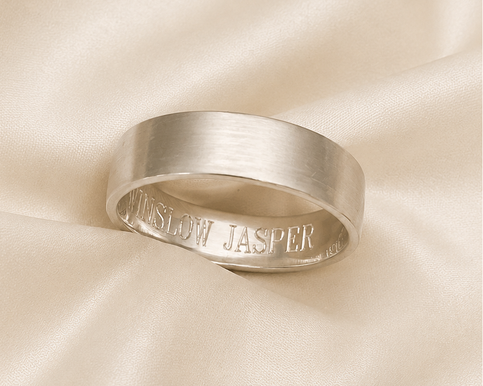 A brushed silver band ring with 'JASPER' engraved on the inside, resting on draped cream satin fabric.