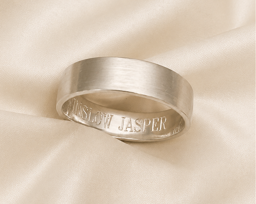 A brushed silver band ring with engraved text reading 'IN SLOW JASPER' resting on draped cream satin fabric.