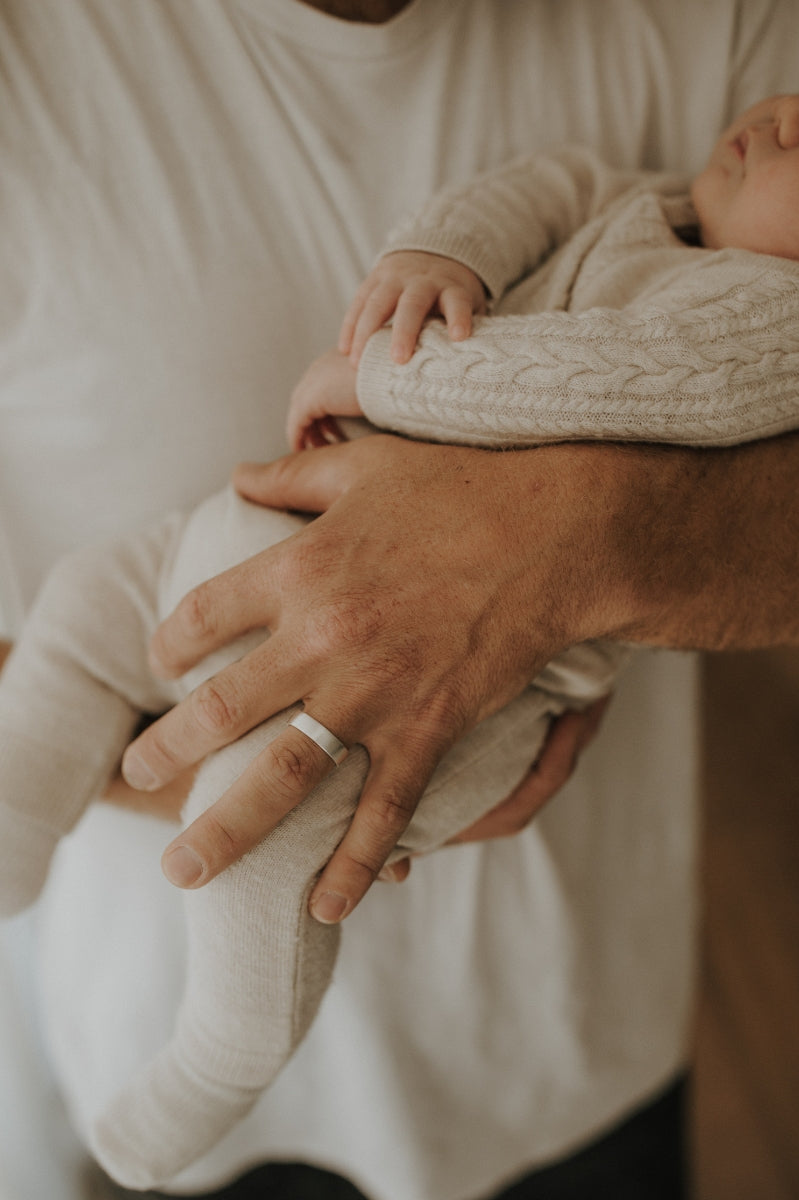 A father wears the Signature Men's Ring on his ring finger. He is holding his newborn son and the ring is being worn on the ring finger of the hand that is holding his son.