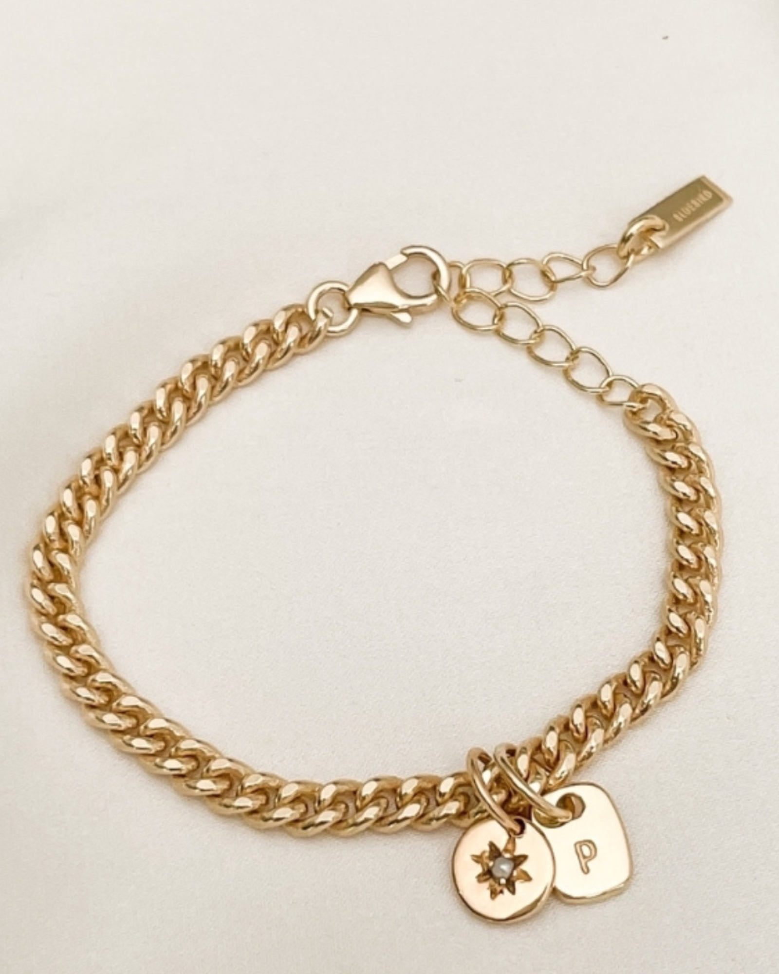 A close-up of a handcrafted gold anklet featuring an engraved rectangular pendant and a daisy pearl charm, designed as timeless personalised jewellery by Bluebird Co