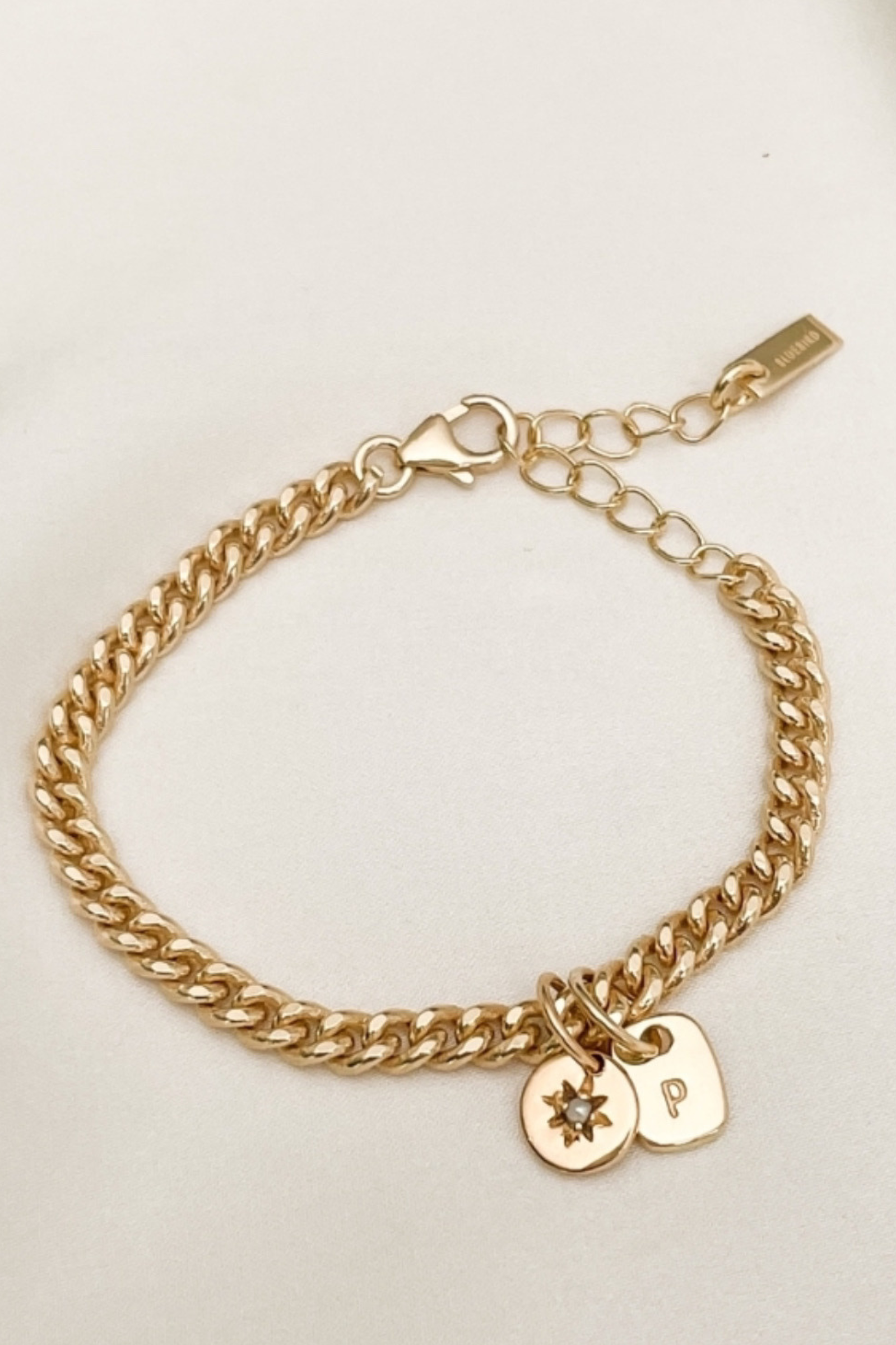 A close-up of a handcrafted gold anklet featuring an engraved rectangular pendant and a daisy pearl charm, designed as timeless personalised jewellery by Bluebird Co