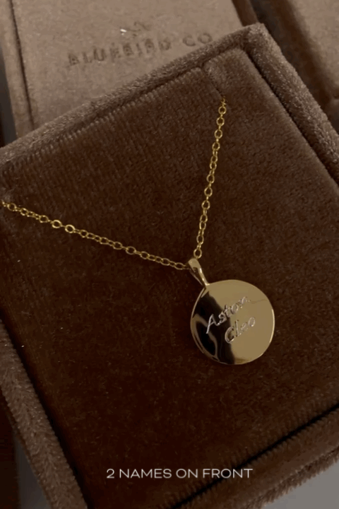 A polished Soleil Necklace in gold with two children's names engraved on the front of it. It is shown in Bluebird Co's signature brown velvet box.