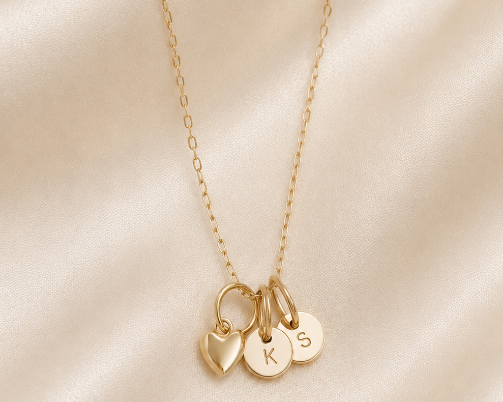 A delicate gold chain necklace with three small hanging charms: a heart, a round initial plate engraved with 'K', and a round initial plate engraved with 'S', displayed on draped champagne satin fabric.