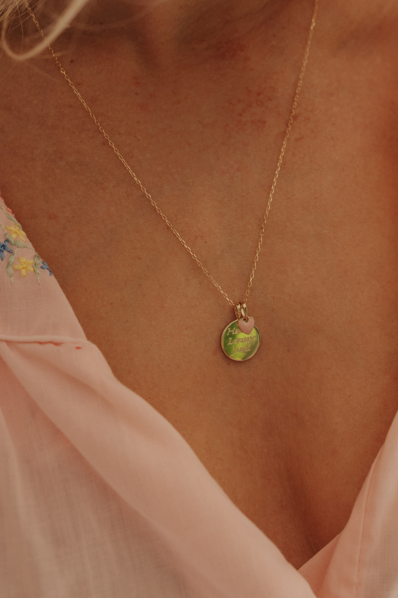 A close up photo of a woman wearing Soléil Pendant in 9ct gold, featuring its signature round, radiant form and personalised engraving with a pink darling pendant.