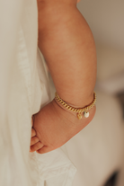 A baby wearing a delicate gold Cuban Link Chain Anklet with a heart charm and personalised pendant, designed as a timeless keepsake piece