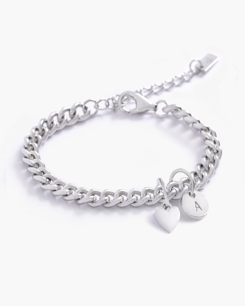A silver Sweetheart Cuban Link Chain Bracelet adorned with a heart charm and engraved initial pendant, designed as a timeless personalised keepsake by Bluebird Jewellery