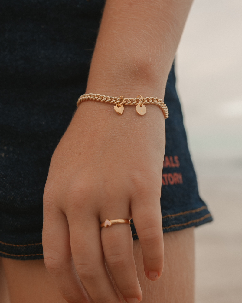 A detailed image capturing a young child wearing a Cuban Link Chain Bracelet with heart charm and initial pendant and matching signet ring, designed as timeless jewellery pieces by Bluebird Co
