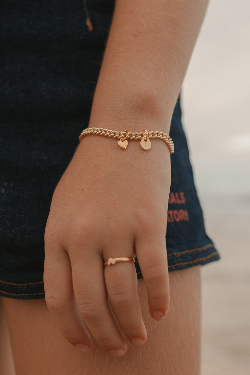 A detailed image capturing a young child wearing a Cuban Link Chain Bracelet with heart charm and initial pendant and matching signet ring, designed as timeless jewellery pieces by Bluebird Co