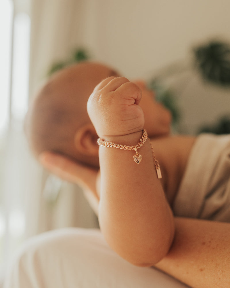 A close up image of a baby wrist wearing a Sweetheart Cuban Link Chain and polished heart pendant, designed as a timeless keepsake piece by Bluebird Jewellery