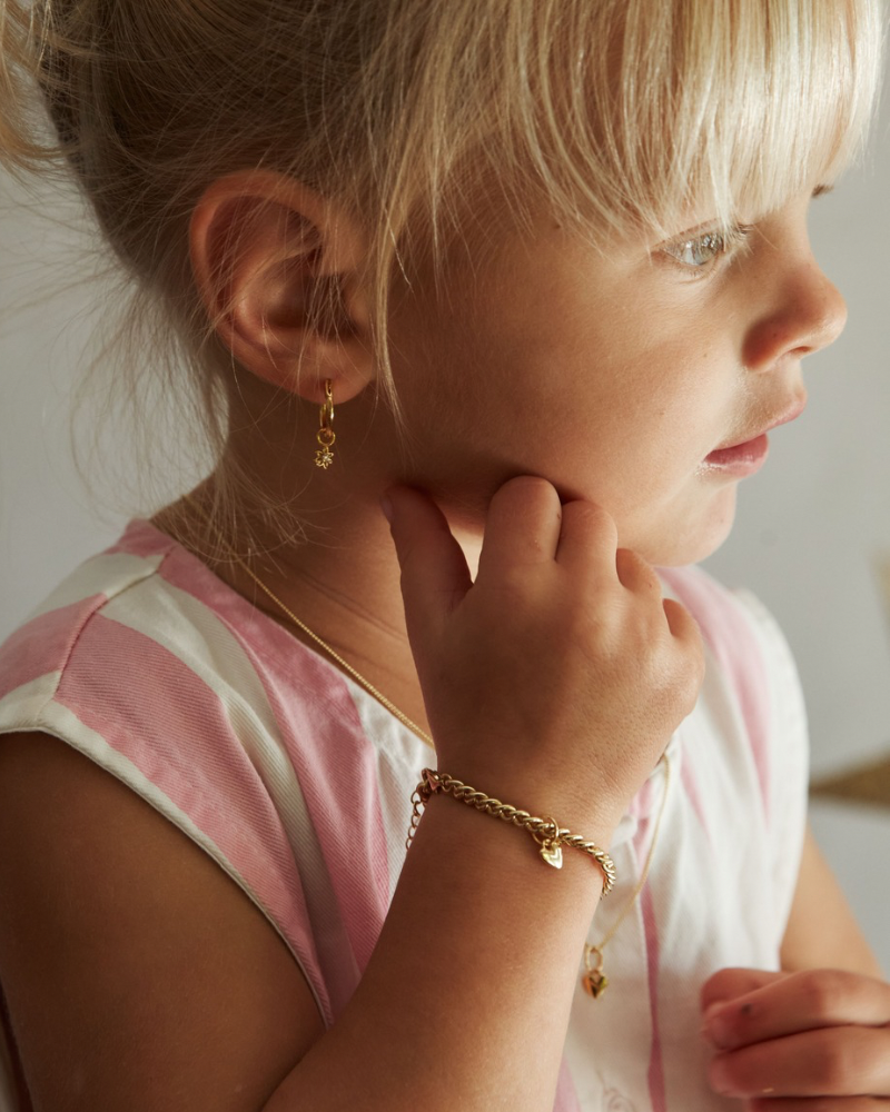 A close-up lifestyle shot of a young girl with blonde hair, wearing a delicate gold chain bracelet with a small heart charm and a matching gold hoop earring, wearing a pink and white striped top.