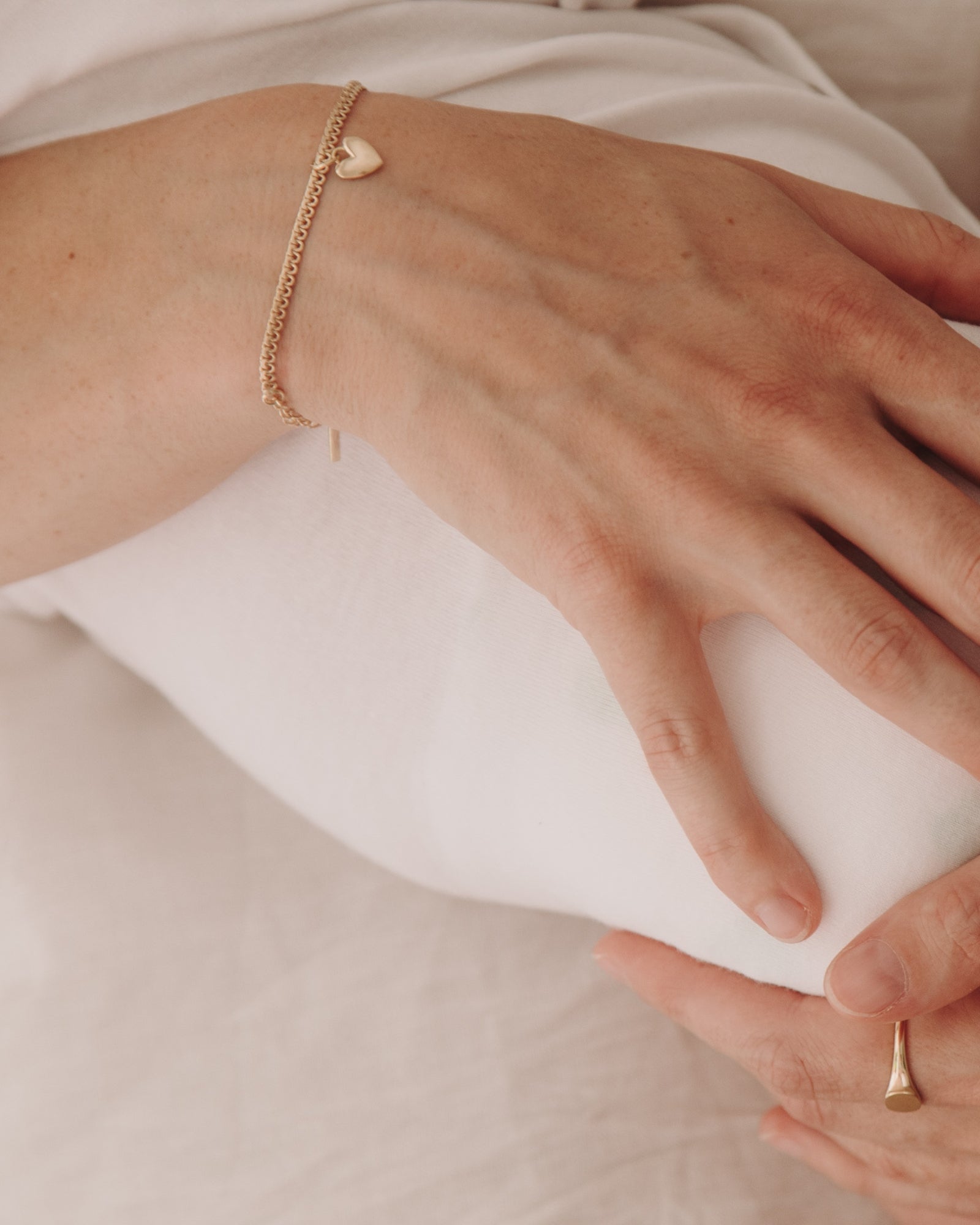 A close-up image capturing a woman holding a baby wearing a Lace Chain Bracelet with heart charm and matching gold signet ring, styled with refined simplicity by Bluebird Jewellery