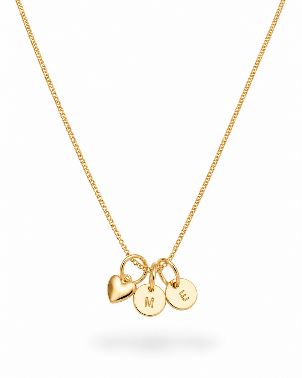 A delicate gold chain necklace featuring three small hanging charms: a heart, a disc engraved with the letter 'M', and a disc engraved with the letter 'E', shown on a clean white background.