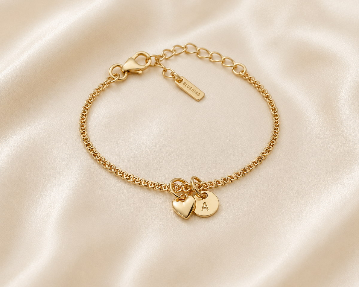A delicate gold chain bracelet with a lobster clasp, adjustable extender chain, and two small heart charms, one engraved with the letter 'A', displayed on a cream satin fabric background.
