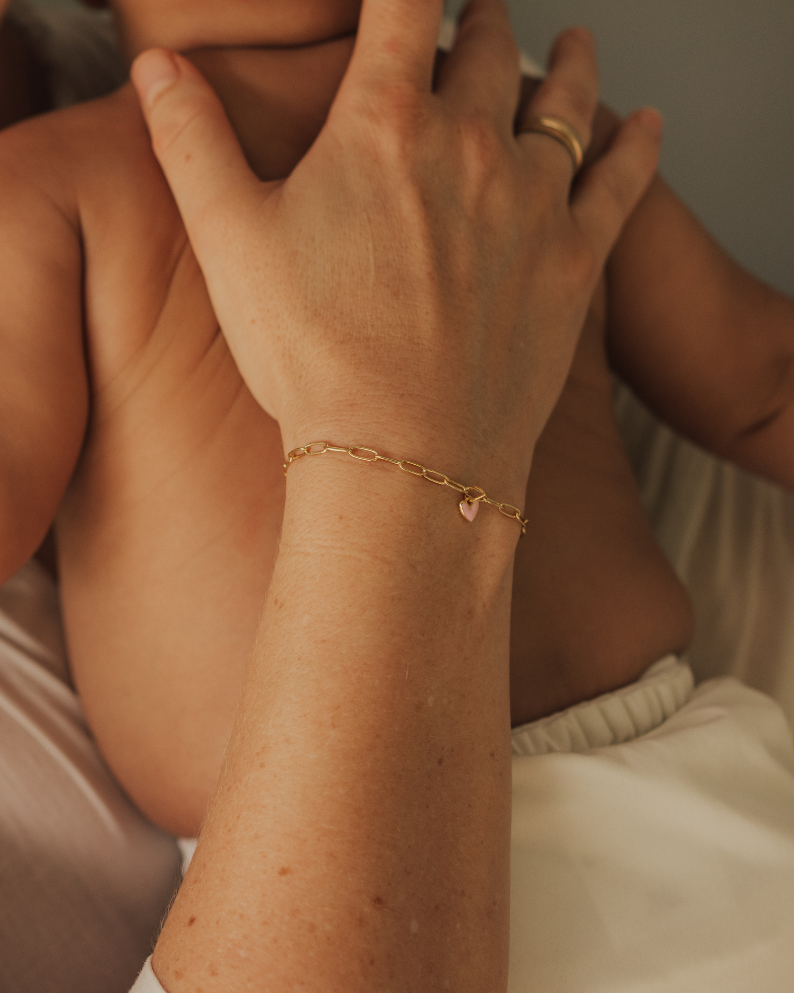 Adult wearing Darling Posie Bracelet Gold with pink heart charm while cuddling baby, a timeless jewellery keepsake by Bluebird Jewellery.