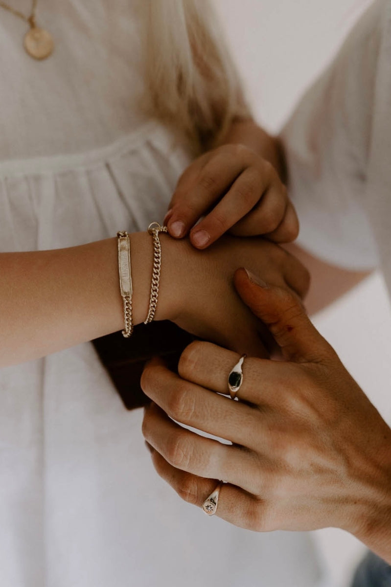 Child wearing engraved gold bracelet while adult helps fasten it, both with gold signet rings, meaningful keepsake jewellery.