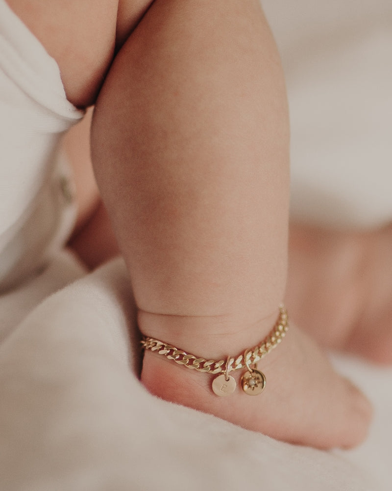 A close up image of a baby's ankle wearing a Cuban Link Chain with a vintage pearl pendant with personalised soild gold initial pendant, designed as a timeless keepsake.