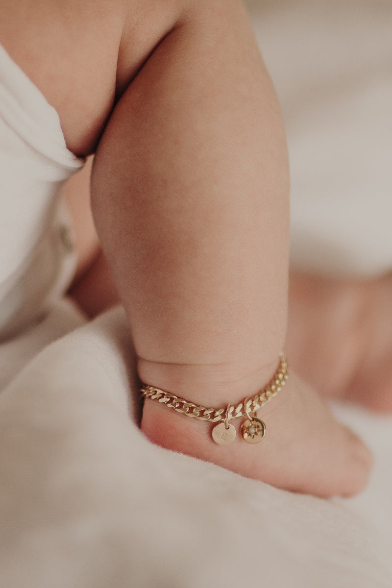 A close up image of a baby's ankle wearing a Cuban Link Chain with a vintage pearl pendant with personalised soild gold initial pendant, designed as a timeless keepsake.