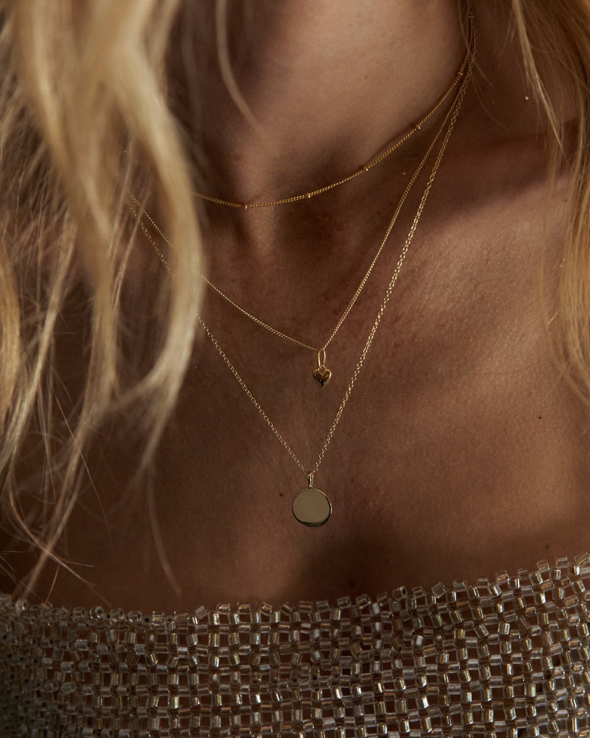 Close-up of a model wearing layered gold necklaces, including a thin chain, a chain with a small heart pendant, and a longer chain with a flat circular disc pendant, paired with a sparkly metallic top.
