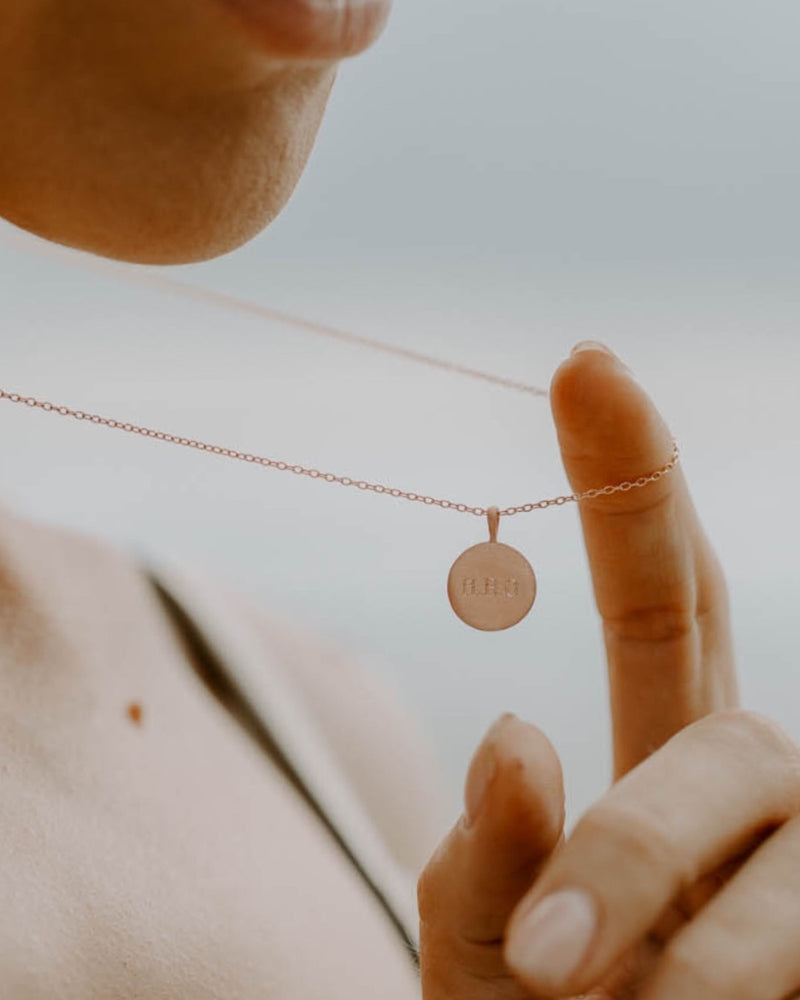 Model holding Petite Signature Necklace in rose gold by the chain to showcase the engraved round pendant in natural light.