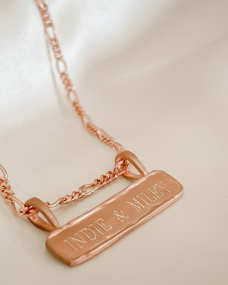 Engraved Signature Bar Necklace in rose gold featuring names. Elegant flatlay of custom pendant with figaro chain.