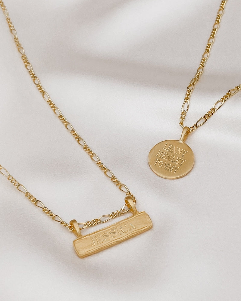 Flatlay of gold Signature Necklace and Bar Necklace with engraved names on soft satin backdrop, showcasing timeless design.