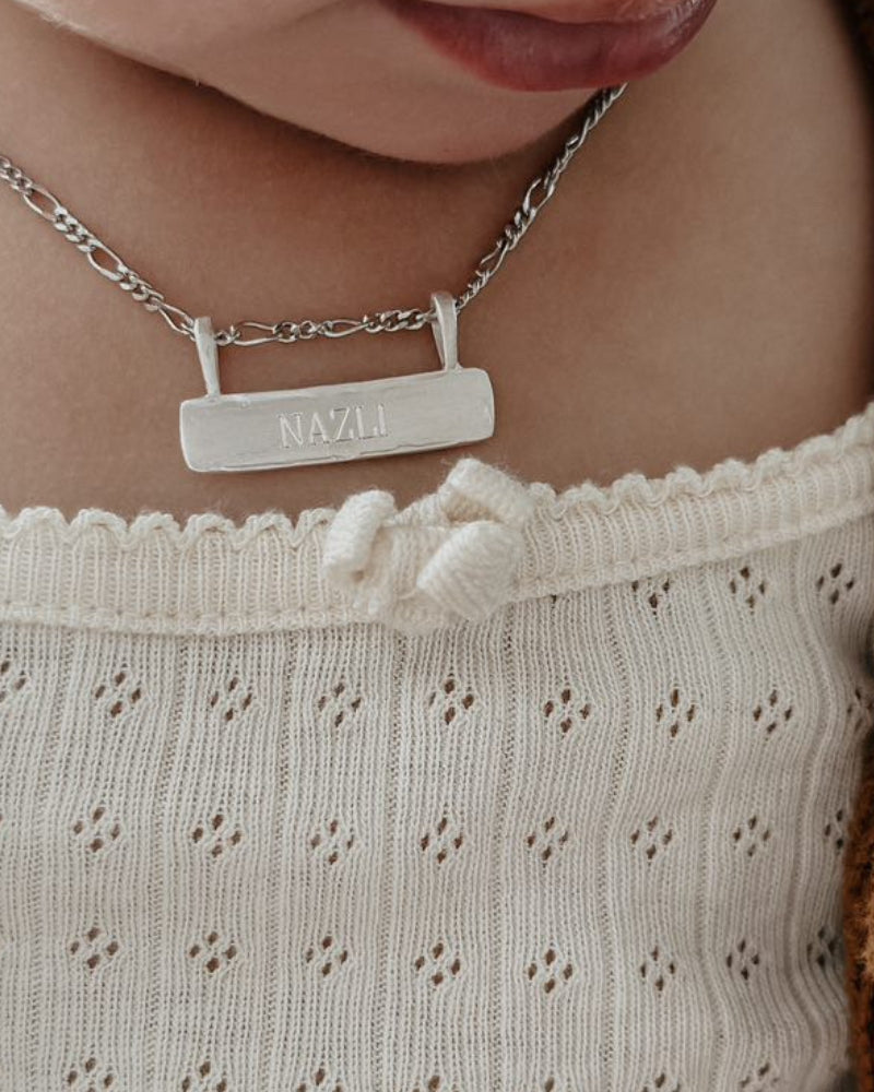 Signature Bar Necklace Silver engraved with “NAZLI” worn by a baby. A sentimental silver piece to mark milestones and memories.