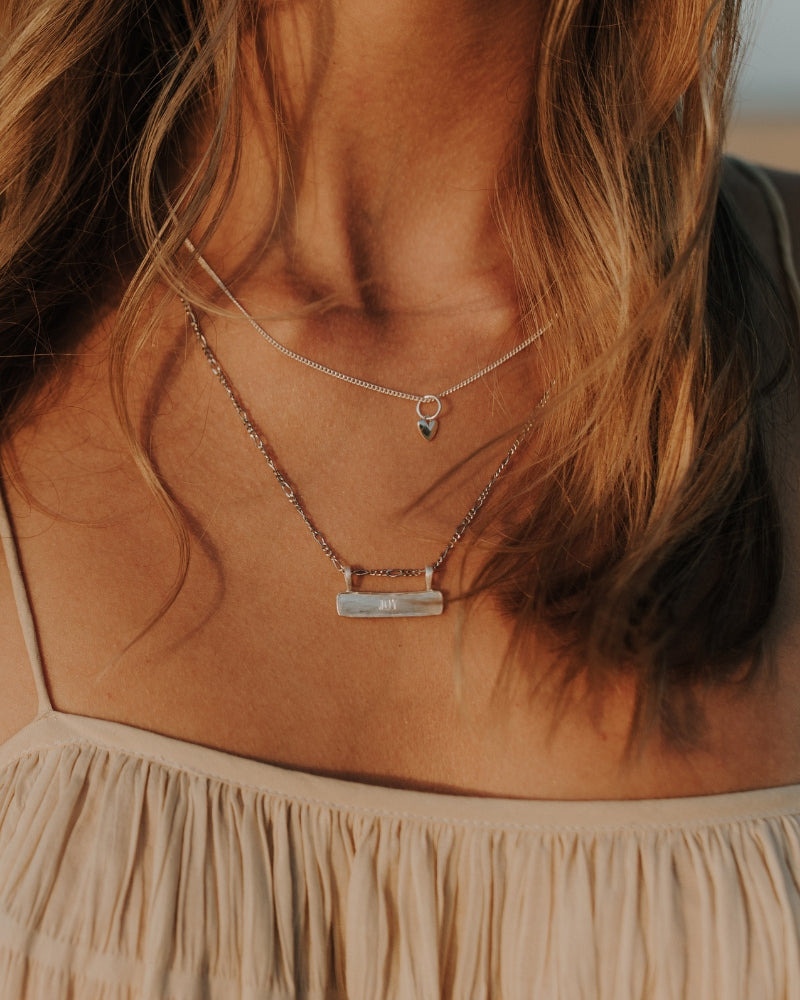 Signature Bar Necklace Silver engraved with “JOY”, layered with a fine heart charm necklace. A delicate and meaningful sterling silver piece.