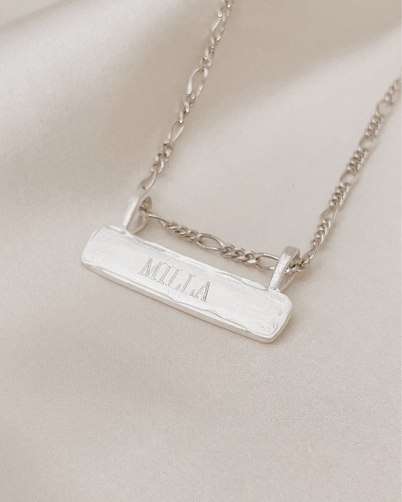 Signature Bar Necklace Silver engraved with “MILLA”, styled on soft fabric to highlight its handcrafted texture and premium sterling finish.