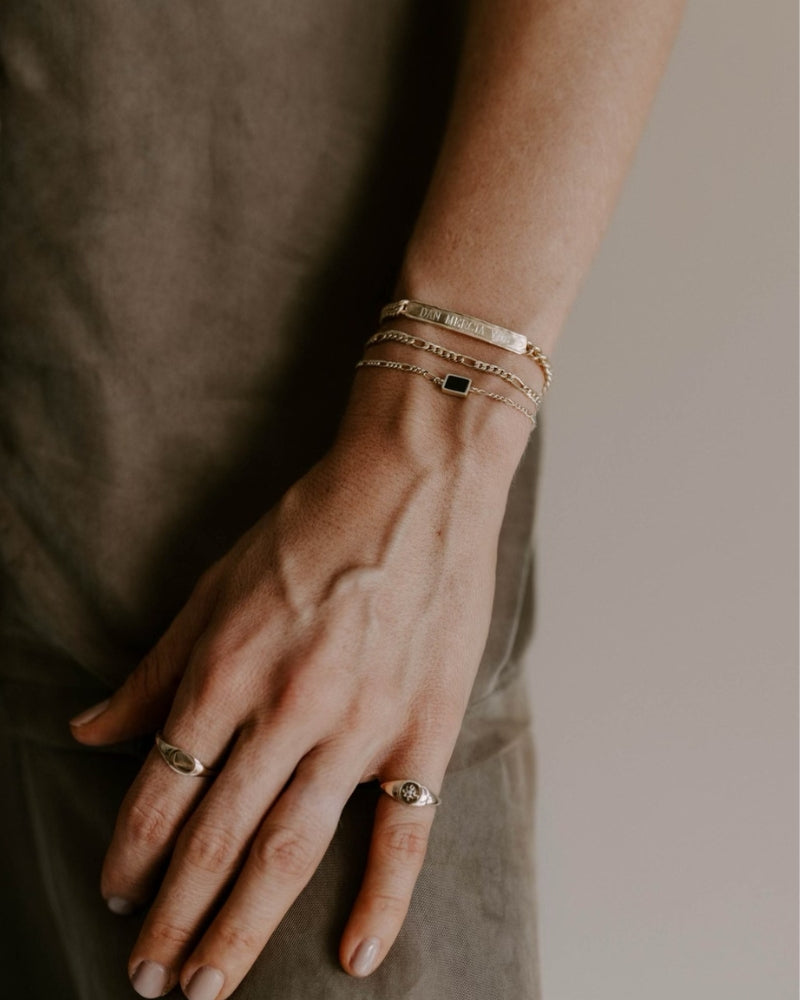 A woman wears the Signature Bracelet engraved with three names, layered with the Figaro and Loved Bracelets in gold vermeil. She also wears the personalised solid gold signet ring and Vintage Pearl Signet Ring by Bluebird Jewellery.
