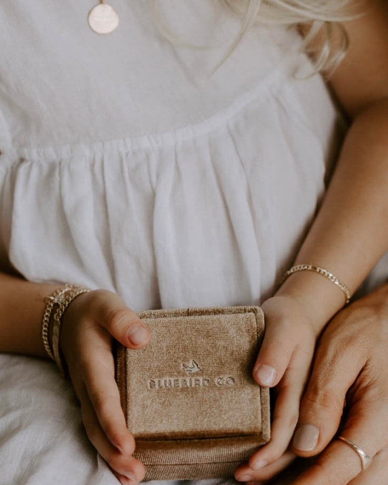 A young girl is holding a velvet gift box with Bluebird Jewellery logo shown close to her Bluebird Co bracelets and personalised necklace.