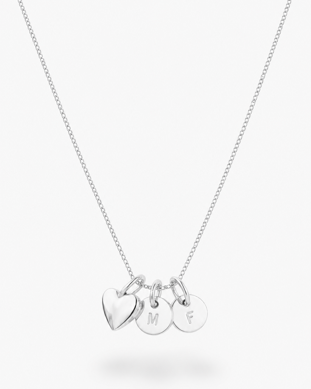 A silver chain necklace with three small hanging charms: a heart-shaped charm and two round disc charms engraved with the letters M, F, and S, shown on a light gray background.