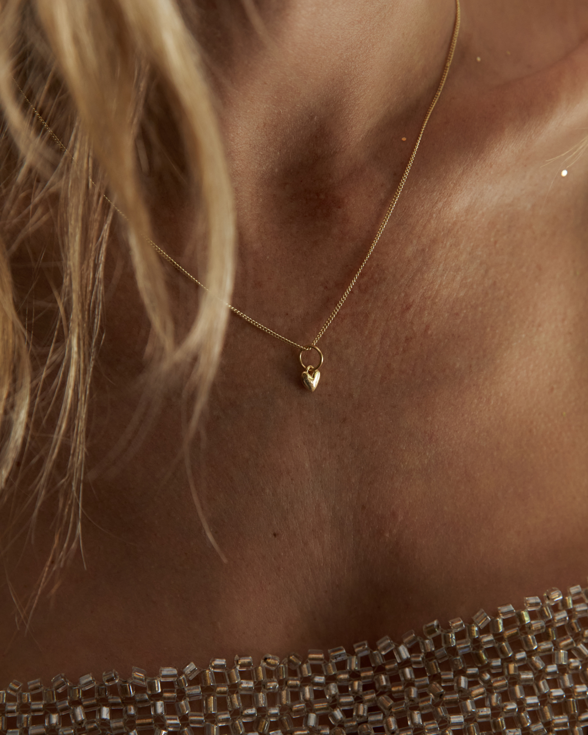A close-up lifestyle shot of a model wearing a delicate gold chain necklace with a small heart-shaped pendant, paired with a sparkling champagne-colored beaded top.