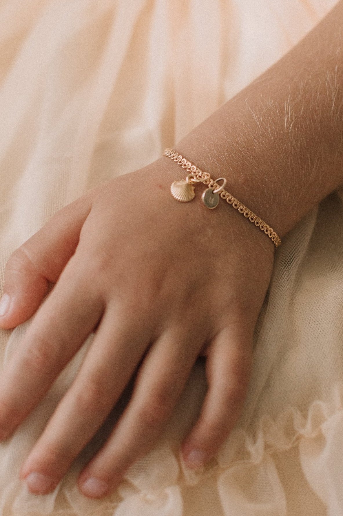 A close-up of a child’s wrist adorned with a gold Lace Chain Anklet with a mermaid shell and soild gold initial pendant, designed as fine personalised jewellery by Bluebird Co