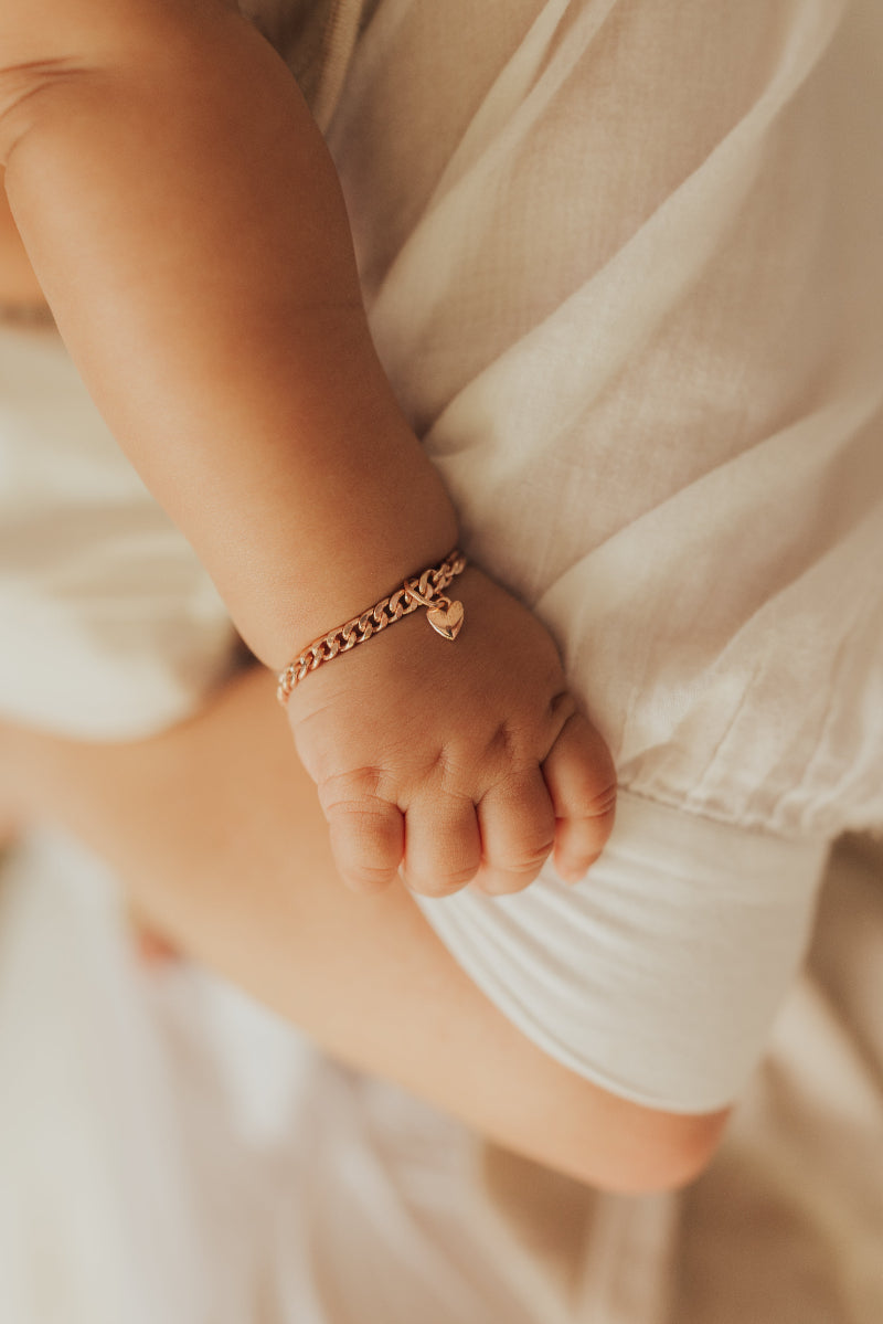 A newborn baby wrist is wearing a Cuban Link Baby bracelet with a petite heart pendant on it by Bluebird Jewellery