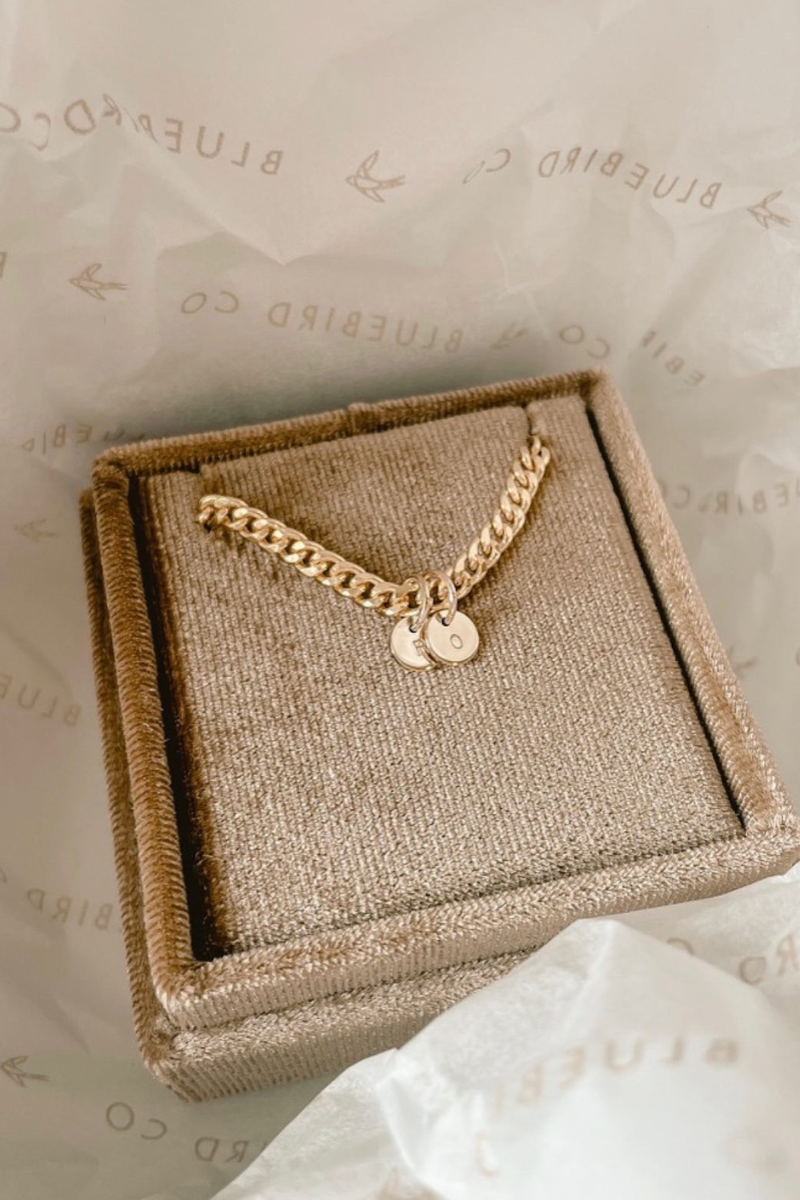 A gold bracelet with personalised pendants, placed inside a Bluebird Co. brown velvet box.