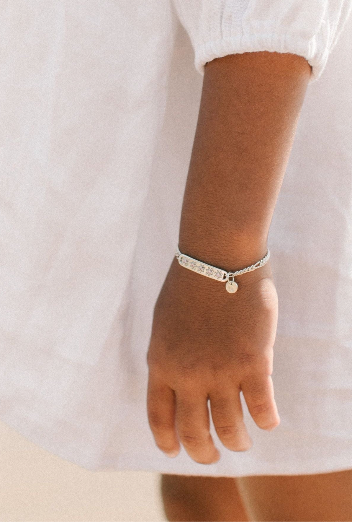 A child wearing a silver Daisy Bracelet by Bluebird Jewellery, featuring five hand-drawn daisies with pink cubic zirconia centres, crafted as a feminine and floral fine jewellery piece along with an initial pendant. 