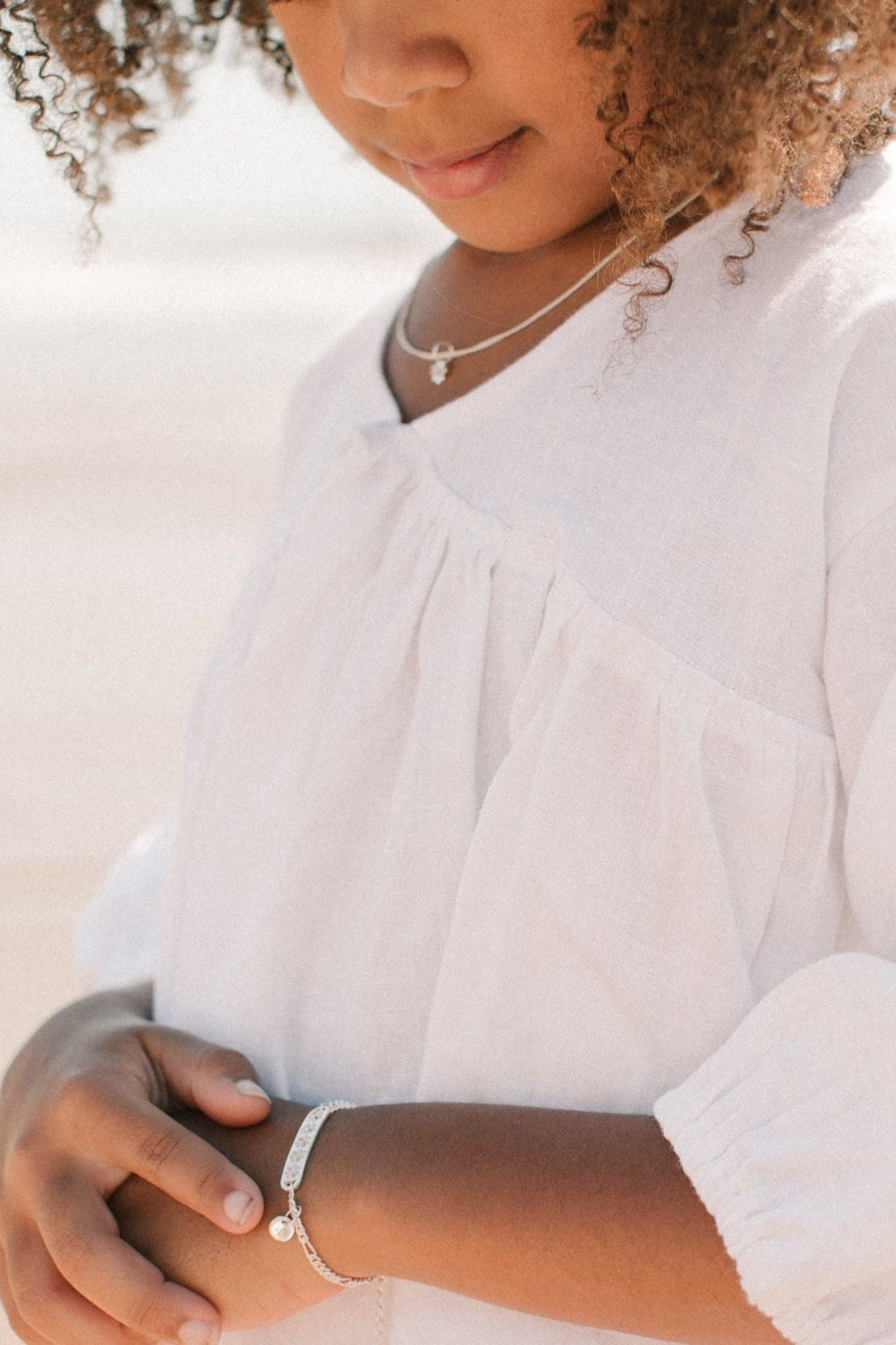 A child wearing a silver Daisy Bracelet by Bluebird Jewellery, featuring five hand-drawn daisies with pink cubic zirconia centres, crafted as a feminine and floral fine jewellery piece along with an initial pendant that represents her name. 