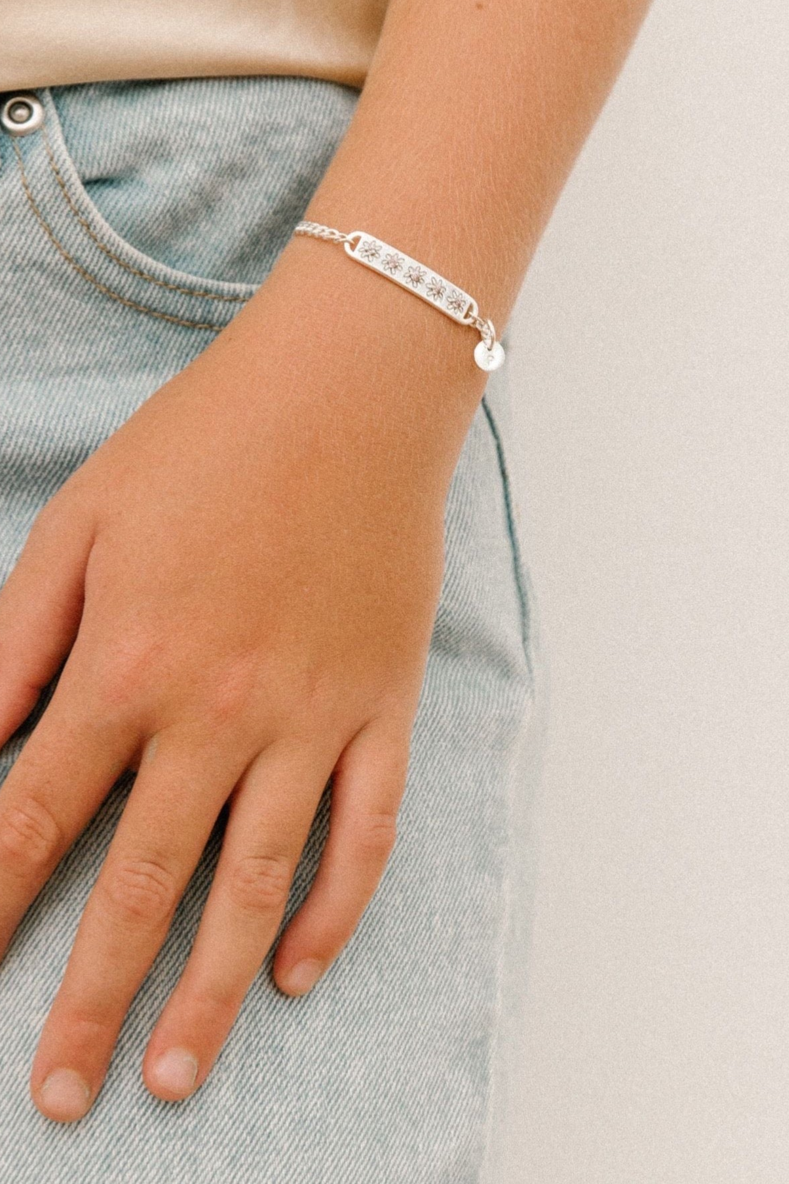 A child wearing a silver Daisy Bracelet by Bluebird Jewellery, featuring five hand-drawn daisies with pink cubic zirconia centres, crafted as a feminine and floral fine jewellery piece along with an initial pendant that represents her name. 