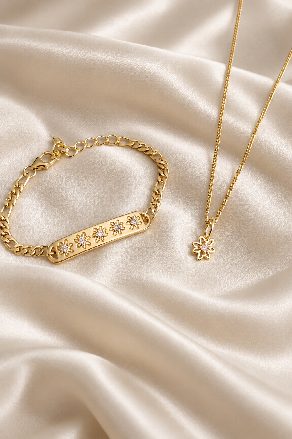 Gold jewelry set featuring a bar bracelet with floral crystal details and a matching flower pendant necklace, displayed on a cream satin fabric background.