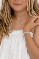 A close-up lifestyle shot of a young girl with blonde hair wearing a delicate silver necklace with a small flower-like pendant and a matching silver bracelet.