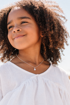 A close-up lifestyle shot of a young girl with curly brown hair wearing a delicate silver necklace with a small flower-shaped pendant, paired with a white top.