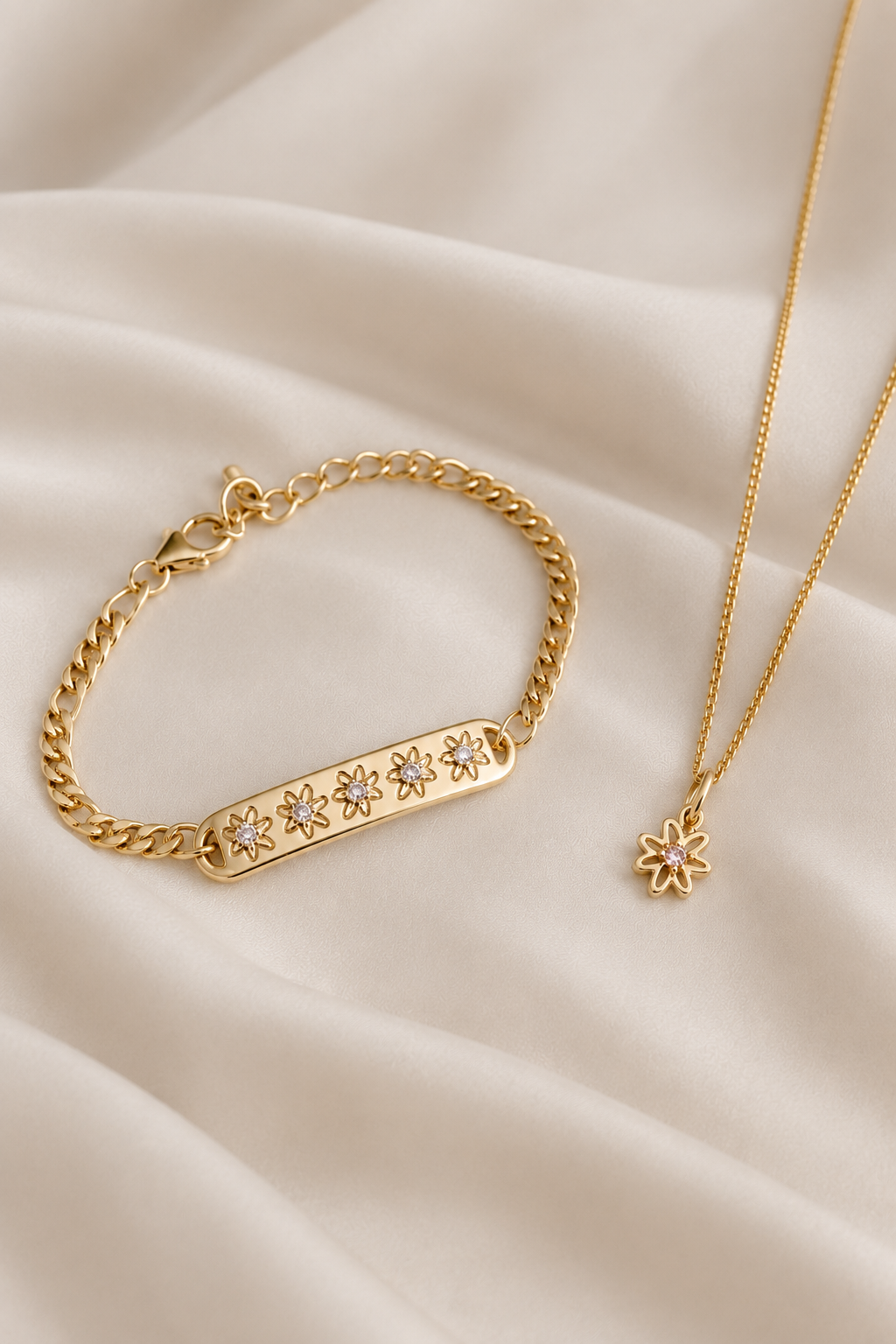 Gold jewelry set featuring a bar bracelet with five star-shaped crystal accents and a delicate chain necklace with a small flower pendant, displayed on draped cream satin fabric.