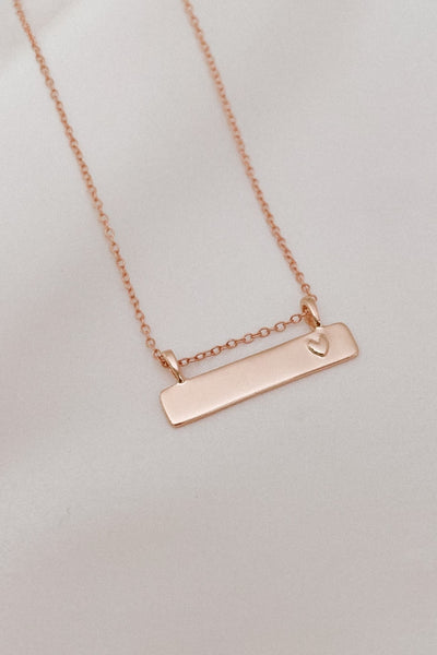 A close up look at the Darling Bar Necklace in  Rose Gold with a smooth, engravable bar pendant with a small heart shape on the right of the pendant.  A sweet Bluebird Necklace that can be personalised for a n adult or a child.