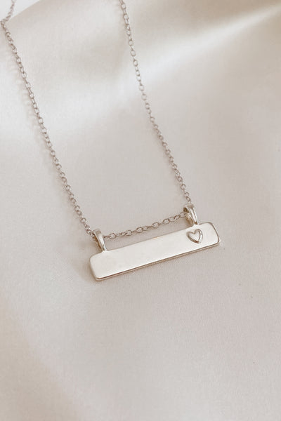 A Darling Bar Necklace rests on a piece of silk.  The beautiful smooth finish on the solid sterling silver bar pendant and petite heart raised on the surface of the front of the bar pendant make this engravable necklace the perfect personalised piece for children and adults.